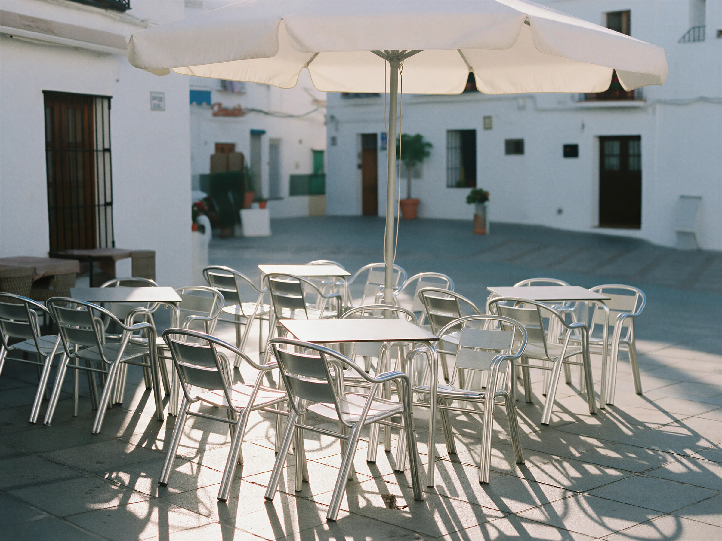 Terraza de bar con mesas y sillas de aluminio y una sombrilla en una plaza encalada