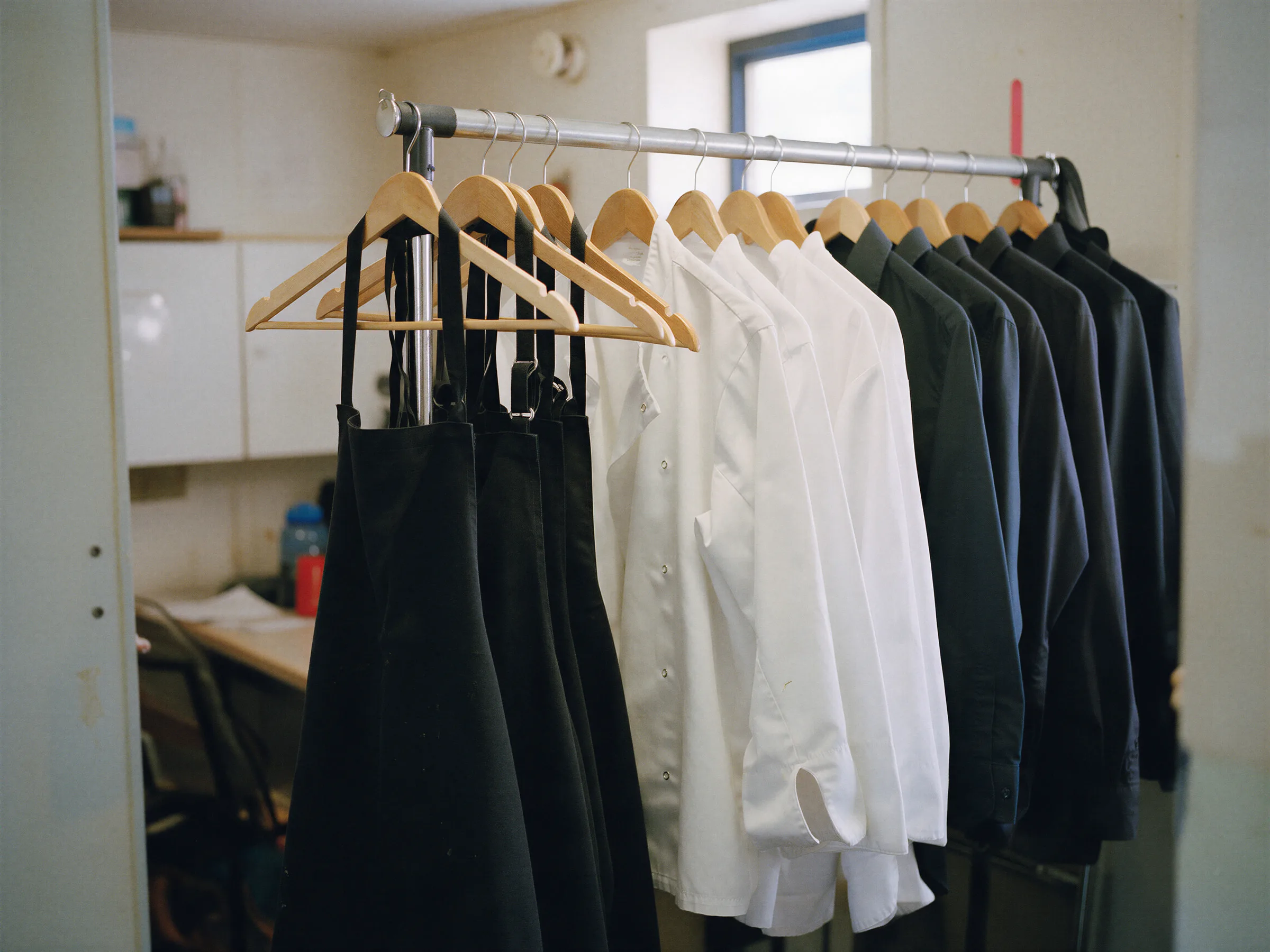 Delantales negros, chaquetillas blancas y camisas oscuras colgados en un perchero
